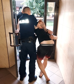 Policjant w umundurowaniu wyprowadza skutego w kajdanki mężczyznę z pomieszczenia.