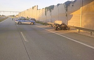 Zdjęcie z wypadku z udziałem motocyklisty. Rozbity motocykl leżący na poboczu, przy barierach energochłonnych, na nim oznakowany radiowóz oraz umundurowani policjanci wykonujący czynności na miejscu zdarzenia.