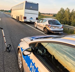 Zdjęcie przedstawia oznakowany radiowóz, a przed nim na poboczu uszkodzony samochód marki peugeot oraz autokar.