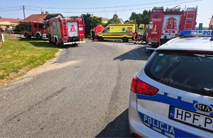 Na zdjęciu widoczny jest oznakowany radiowóz, dwa wozy bojowe straży pożarnej oraz karetka pogotowia. Obok rozłożony jest czerwony parawan z napisem straż.