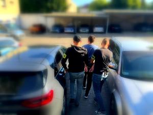 Dwóch policjantów w cywilnych ubraniach z opaska na ręce z napisem policja trzymają mężczyznę z założonymi kajdankami.