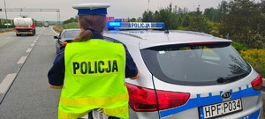 Policjantka, radiowóz i samochód stojący przed radiowozem, inne auta jadące po drodze.