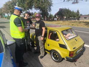 Umundurowany policjant wręczający materiały edukacyjne dwóm mężczyznom podróżującym małym fiatem.