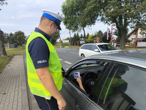 Umundurowany policjant ruchu drogowego wręczający kierującym  naklejki promujące kampanię &quot;Mistrzu Zwolnij&quot;.