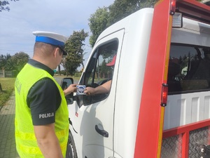 Umundurowany policjant ruchu drogowego wręczający kierującym  naklejki promujące kampanię &quot;Mistrzu Zwolnij&quot;.