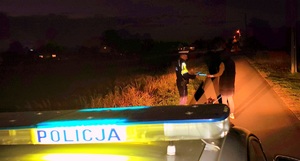 Napis policja na belce radiowozu i policjant wręczający odblask pieszej.