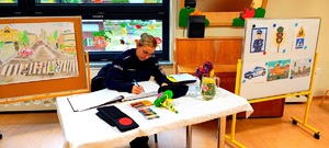 Policjantka wpisująca die do księgi gości, obok rysunki o bezpieczeństwie.