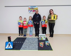 Policjantka, dzieci w kamizelkach odblaskowych trzymające znaki drogowe i nauczycielka na podłodze lezy mata z przejściem dla pieszych.