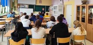 Policjantka mówiąca do młodzieży siedzącej w ławkach szkolnych.