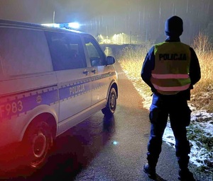 Policjant w kamizelce odblaskowej z napisem policja pora nocna stoi przy radiowozie obok torów.