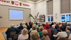 Komendant siedzący przy stole i seniorzy na krzesłach.