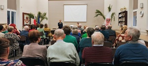 Policjantka mówiąca do mikrofonu i seniorzy na krzesłach.