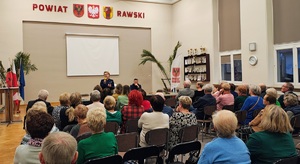 Policjantka mówiąca do mikrofonu i seniorzy na krzesłach.