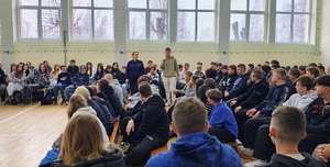 Grupa młodzieży siedząca w ławkach i chłopiec z mikrofonem i policjantka.