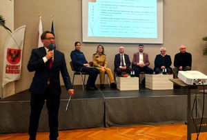 Mężczyzna z mikrofonem w tle przedstawiciele służb siedzący na krzesłach.