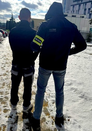 Policjant w cywilnym ubraniu z opaską z napisem policja i zatrzymany stojący obok.