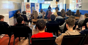 Policjantka oraz psycholog stoją przed dziećmi siedzącymi na krzesłach.