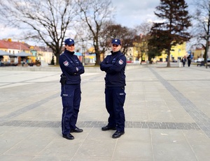 Policjant i policjantka stoją na placu z założonymi rękami.