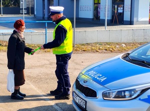 Policjant wręcza element odblaskowy starszej pani, obok radiowóz.