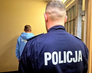 Policjant stojący tyłem, w pomieszczeniu za kratami stoi tyłem zatrzymany.