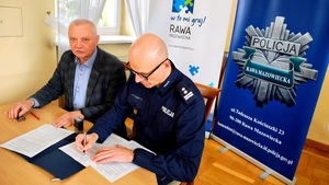 Komendant i zastępca Burmistrza siedzą przy stole i podpisują dokumenty, w tle rolapy instytucji.