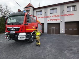 Kobieta w stroju strażackim przy radiowozie i strażnicy.