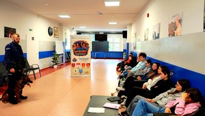 Policjant z psem służbowym i dzieci siedzące na krzesłach.