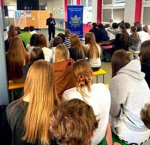 Policjantka stojąca przed grupa młodzieży siedzącej na ławkach.