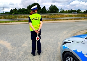 Policjantka z tarczą w ręce do zatrzymywania pojazdów, radiowóz na poboczu.