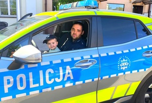 Policjant siedzący w radiowozie z dzieckiem, które ma na głowie czapkę policyjną.