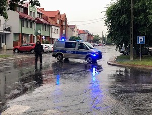 Radiowóz w poprzek drogi i policjant przed autem w deszczu.