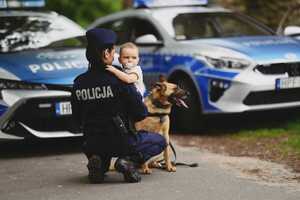 Policjantka z dzieckiem na tle radiowozów i pies służbowy.