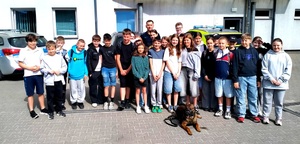 Dzieci stojące z policjantami oraz nauczycielami, a przed nimi leży pies służbowy.