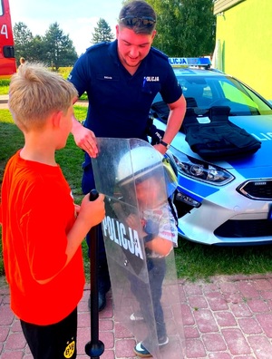 Policjant, radiowóz, dziecko w  tarcza policyjna w kasku oraz inne dziecko obok.